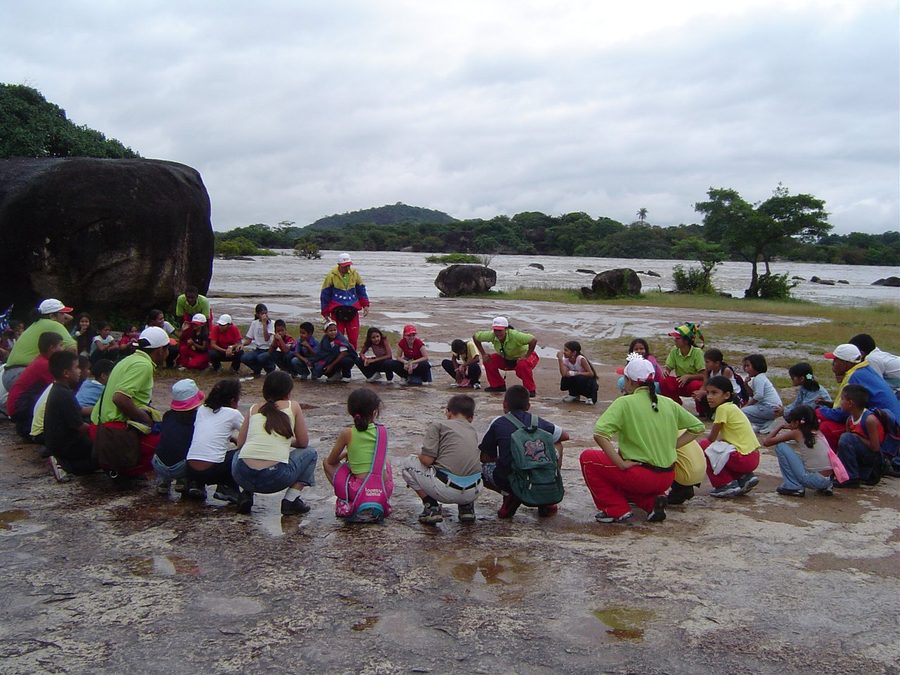 Facilitador de pie en el centro de un amplio círculo de niños sentados sobre una laja de piedra junto a un río, con vegetación y colinas al fondo