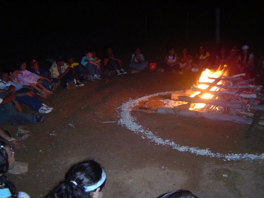 Participantes sentados en círculo alrededor de una gran fogata encendida durante la noche, en el cierre de un campamento