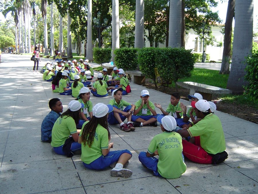 Varios subgrupos de niños uniformados de verde sentados en círculos de conversación sobre una plaza arbolada, acompañados por facilitadores